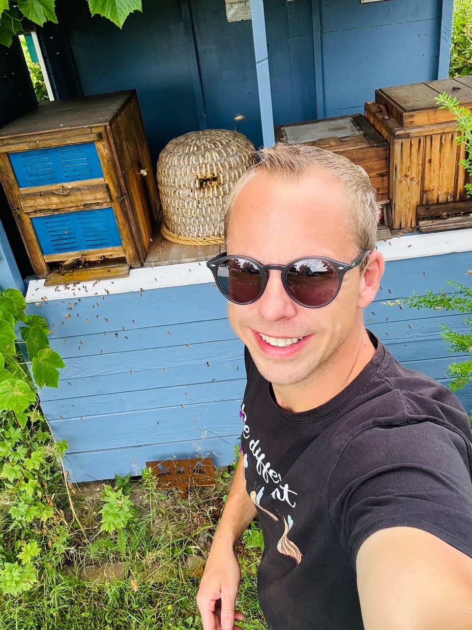 Mann mit Sonnenbrille lächelt vor einer blauen Wand und Bienenbeute in der Natur.