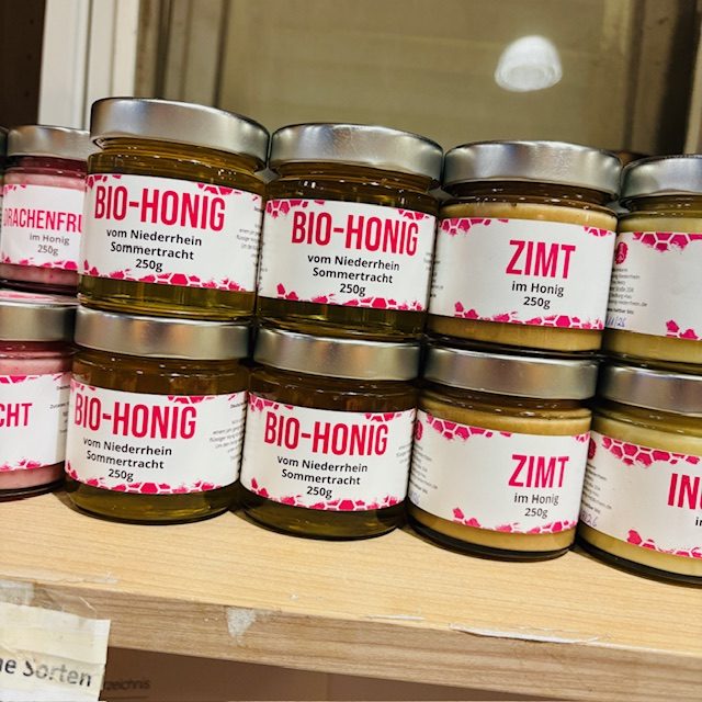 Sechs Gläser mit Bio-Honig in unterschiedlichen Farben auf einem Regal.