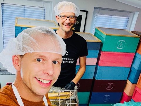 Zwei Männer in Haarnetzen lächeln vor bunten Boxen in einem Raum.