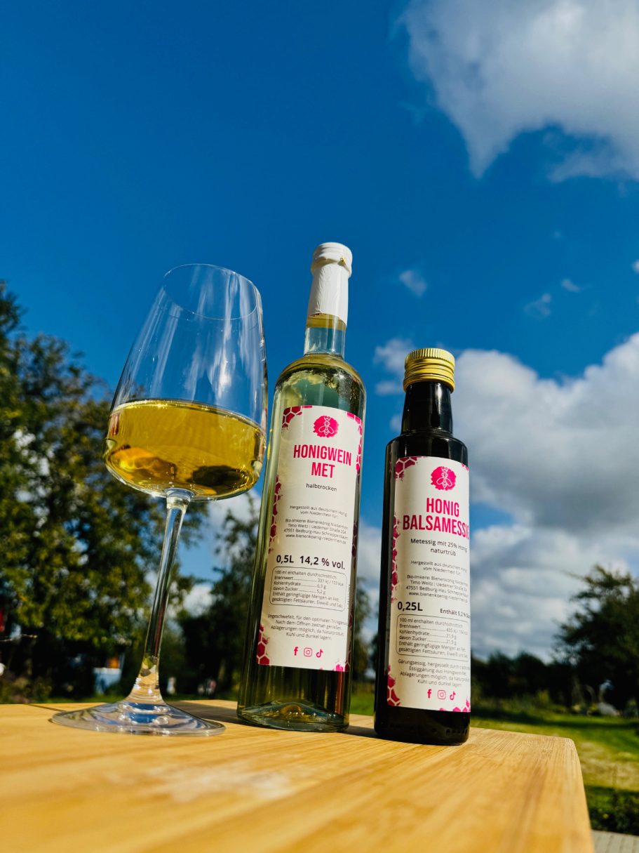 Weißwein und Öl in Flaschen neben einem Weinglas auf einem Holztisch, blauer Himmel im Hintergrund.