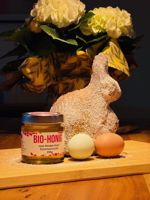 Osterhase aus Teig, zwei Eier und ein Glas Marmelade vor einer Blumenarrangement.