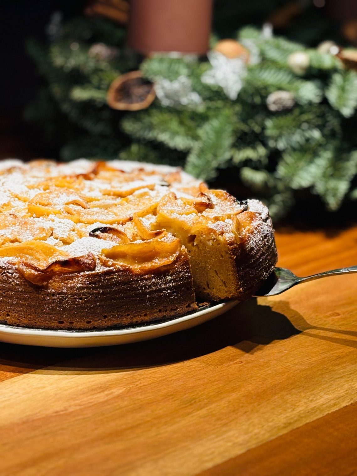 Apfelkuchen mit Puderzucker auf einem Holzbrett, dekoriert mit Weihnachtskränzen.