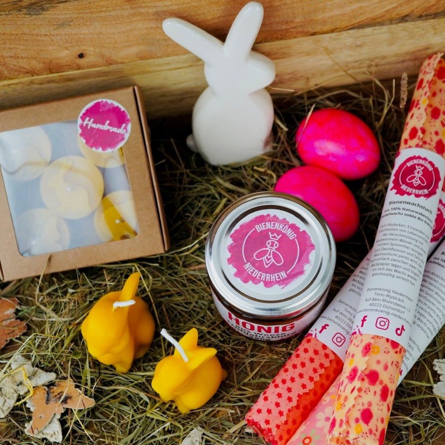 Geschenkset mit bunten Ostereiern, Kerzen, Marmelade und einem Hasen in Heu.