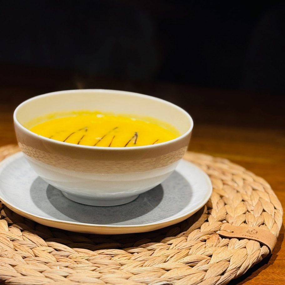 Eine Schüssel mit dampfender, orangefarbener Suppe auf einem Untersetzer.