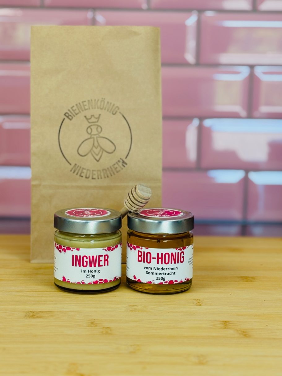 Zwei Gläser Honig mit bunten Etiketten vor einer gemusterten Wand und einer braunen Tüte.