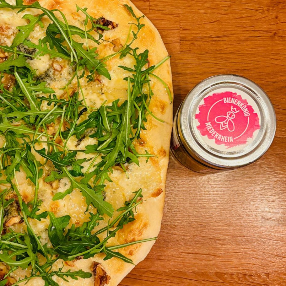 Pizza mit Rucola neben einem Glas mit pinkem Deckel.