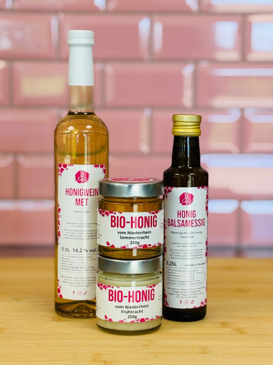 Vier Flaschen mit Lebensmitteln: Essig, Öl und Gewürze auf einem Holzbrett vor einer rosafarbenen Wand.